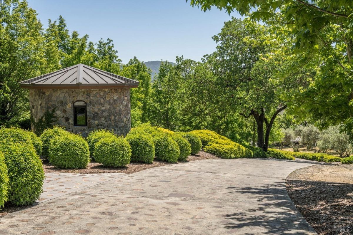 a small round stone building with a metal roof, surrounded by well-manicured green shrubs and trees. A paved pathway leads alongside the structure and curves gently into the distance. In the background, there are lush trees and rolling hills under a clear blue sky.