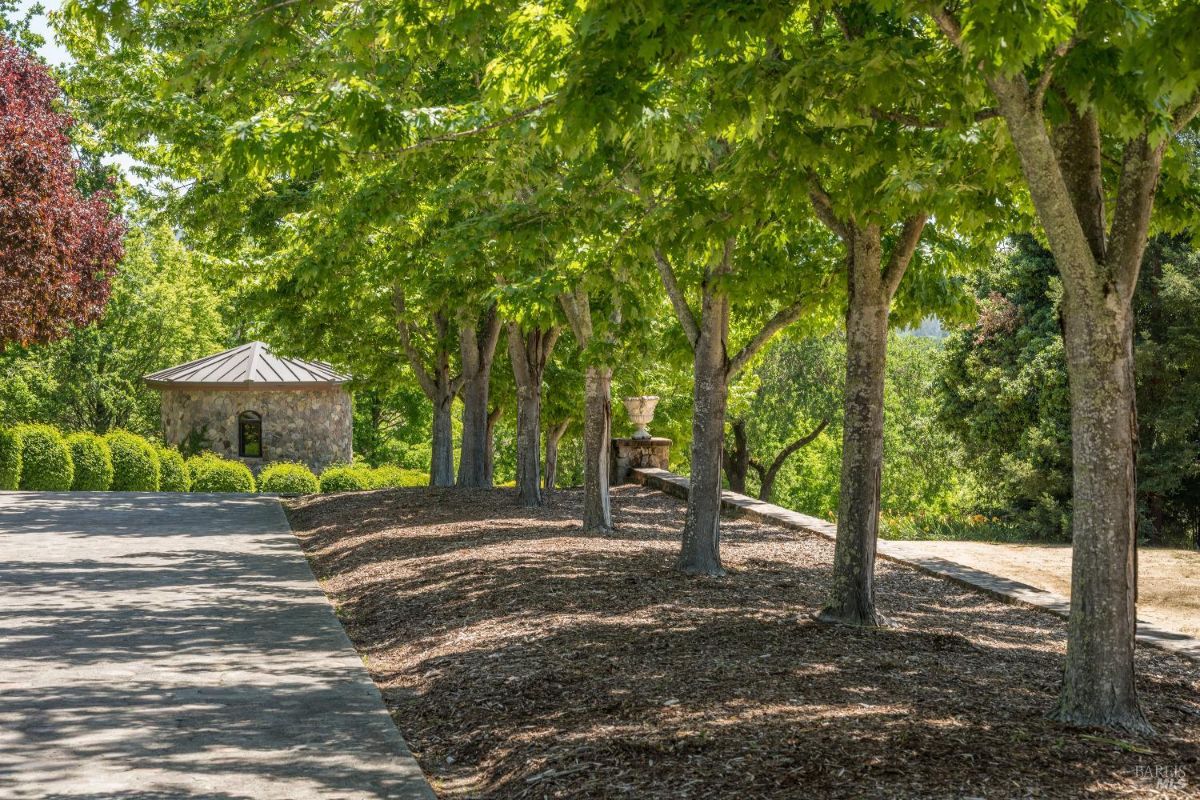 a small round stone building with a metal roof, surrounded by well-manicured green shrubs and trees. A paved pathway leads alongside the structure and curves gently into the distance. In the background, there are lush trees and rolling hills under a clear blue sky.