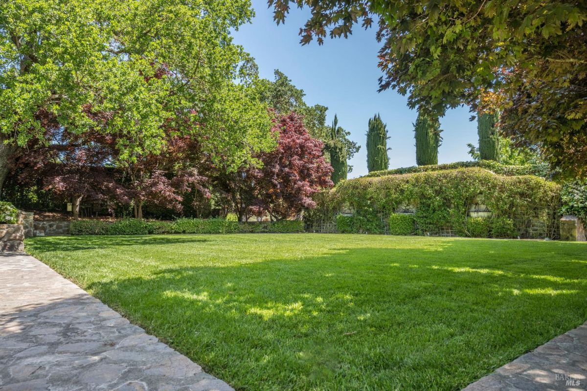 Well-maintained green lawn surrounded by mature trees, colorful foliage, and trimmed hedges. A stone pathway borders the lawn on the left side, leading into the landscaped area. The scene is framed by a variety of greenery and tall cypress trees in the background.