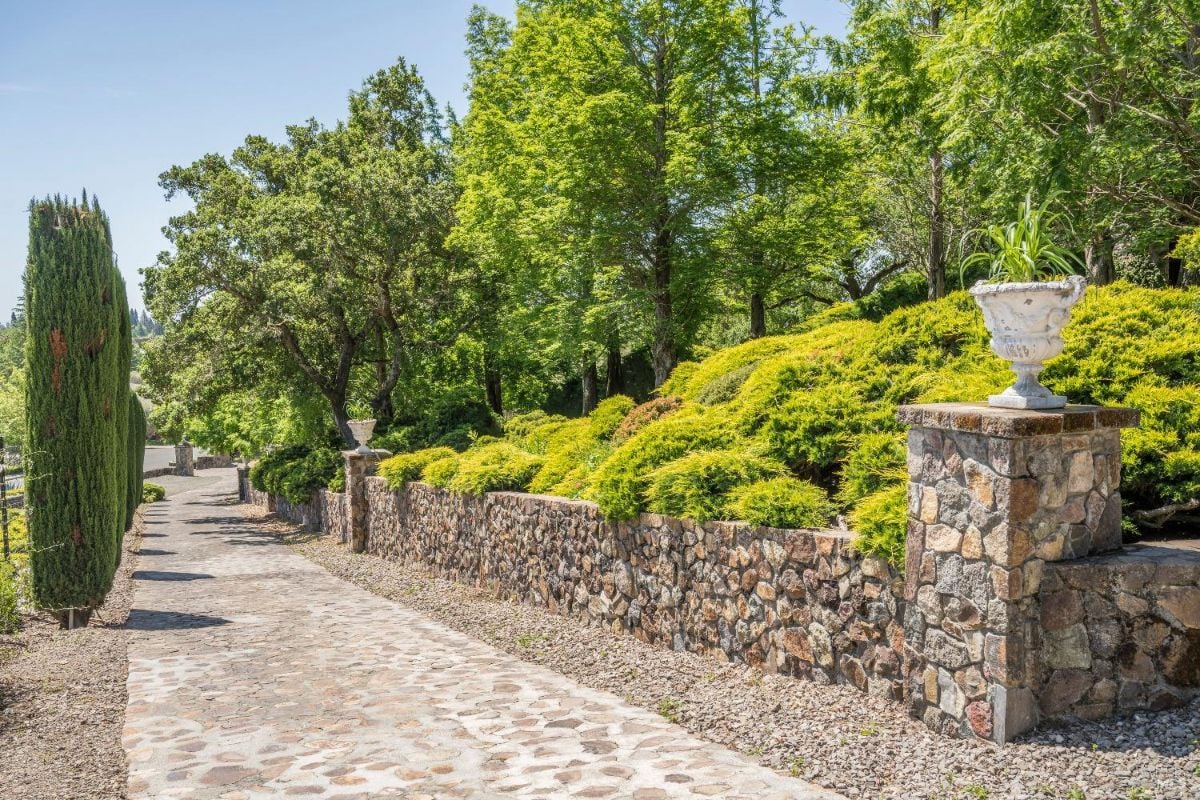 Landscaped garden pathway lined with a stone wall and lush greenery. The path is paved with stones, bordered by tall cypress trees on the left and trimmed bushes on the right. Decorative stone pillars with potted plants stand along the wall.