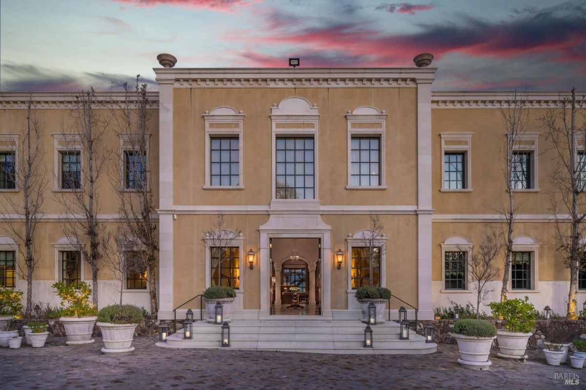 Exterior of a two-story building with a symmetrical design, large rectangular windows, and a central entrance. The facade is beige with white decorative trim, including columns, window borders, and a detailed cornice. In front of the entrance, there are steps leading to a pair of doors, surrounded by potted plants and lanterns, with a cobblestone courtyard visible in the foreground.
