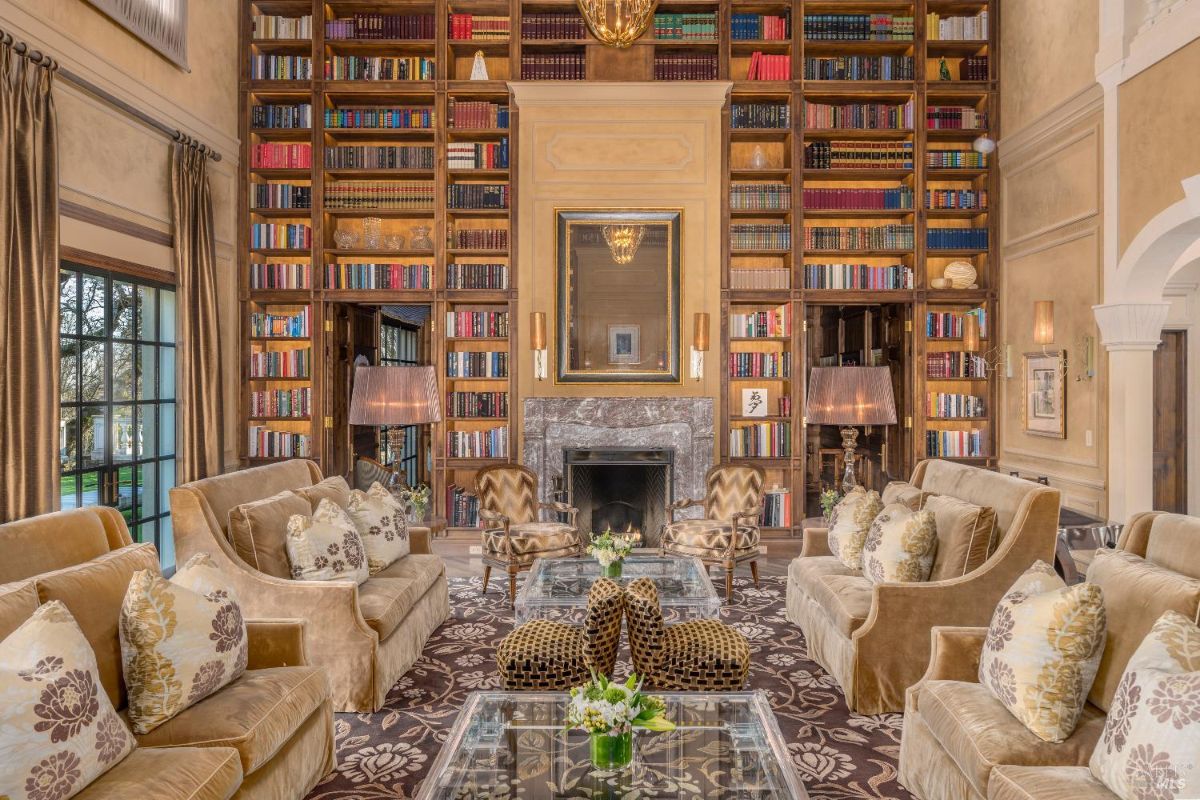 Large living room with a floor-to-ceiling bookshelf covering the entire back wall, filled with colorful books. In the center, there is a fireplace with a marble surround and a mirror above it, flanked by two tall lamps. The seating area includes plush armchairs, sofas, and glass-topped coffee tables arranged on a patterned rug, with decorative pillows and small floral arrangements on display.