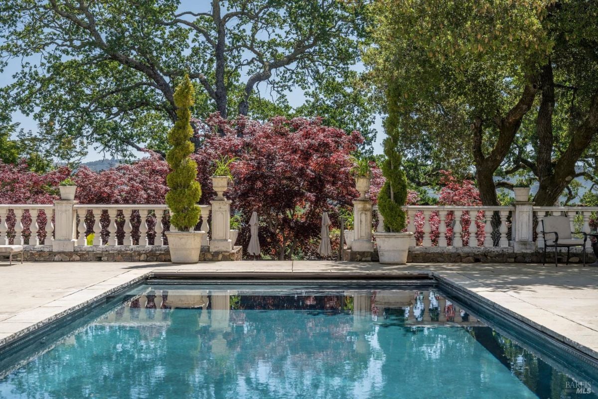 Rectangular swimming pool surrounded by a paved deck area, reflecting the surrounding trees and sky. Ornamental white balustrades line the perimeter, accompanied by potted plants and two neatly trimmed topiary trees. Behind the balustrade, there are red-leafed trees, green foliage, and large oak trees providing a natural backdrop.