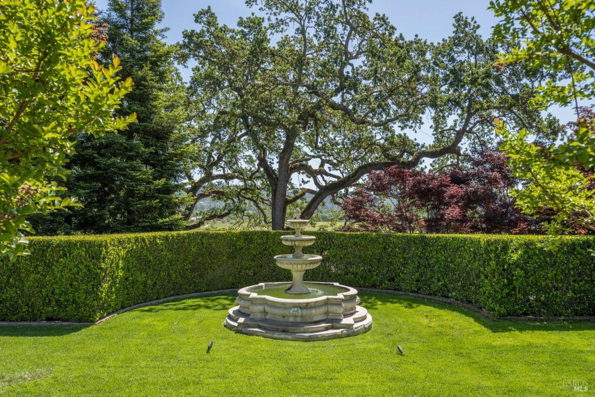 A tiered stone fountain situated in the center of a well-manicured lawn. Behind the fountain, a neatly trimmed hedge creates a boundary, with large mature trees, including red-leafed and green foliage, in the background. The area is surrounded by lush greenery under clear blue skies.