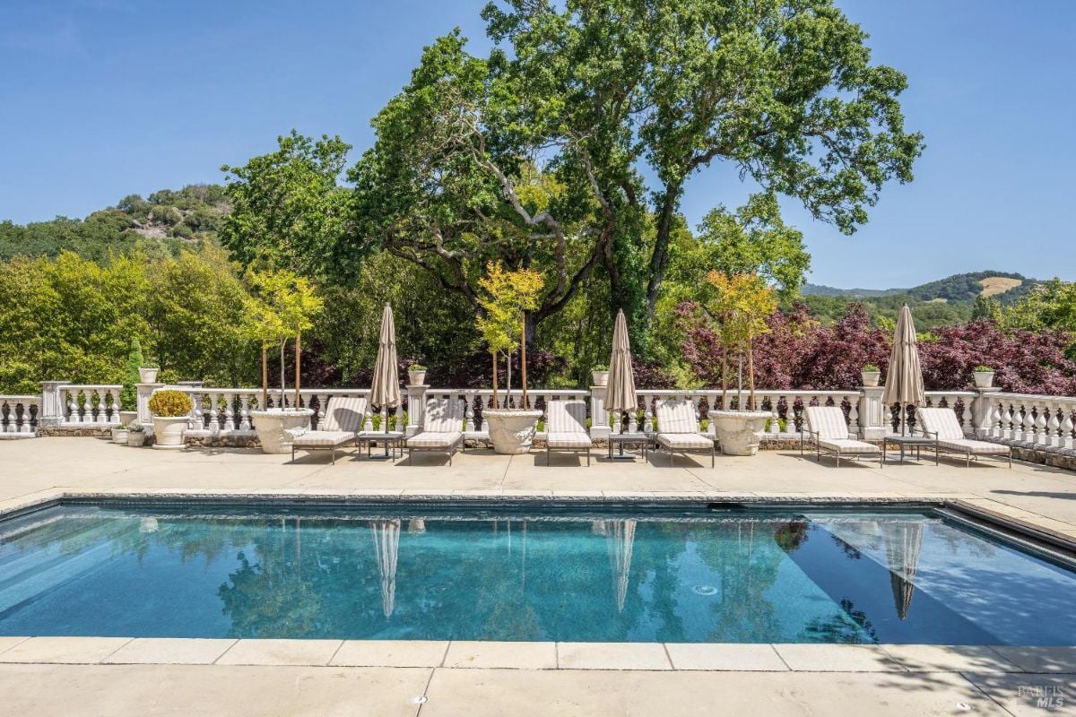 Pool surrounded by a stone deck with multiple lounge chairs and umbrellas. Potted plants and balustrades line the perimeter of the pool area, with tall trees and rolling hills in the background. The reflection of trees and umbrellas is visible on the surface of the calm pool water.