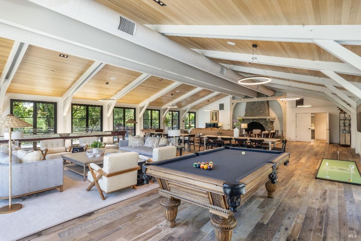 Spacious recreational area with a pool table in the foreground and a small putting green to the side. Seating areas with sofas, chairs, and coffee tables are arranged on a large rug, facing windows that provide ample natural light. A stone fireplace is located at the far end of the room, surrounded by additional tables and chairs for dining or activities.