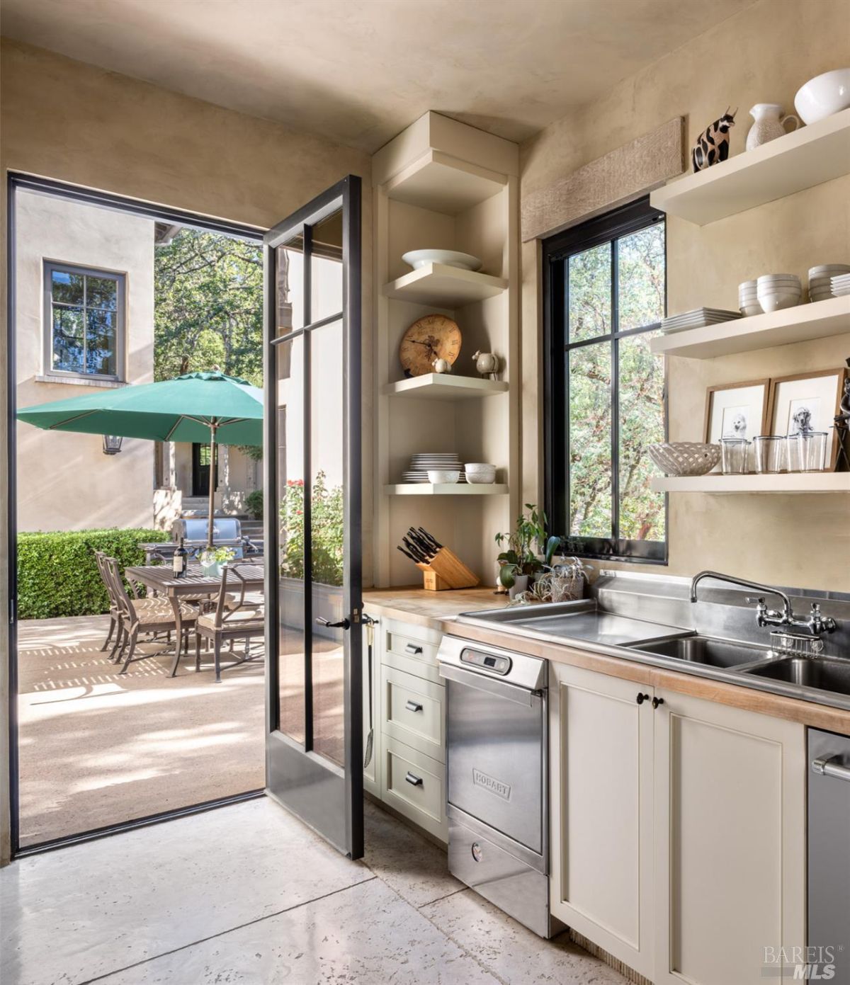 This compact kitchen opens up to an outdoor patio through French doors, letting in natural light. It features a farmhouse sink, minimalist shelving, and a mix of neutral tones. Outside, a patio with green umbrellas adds a charming connection to the outdoors.