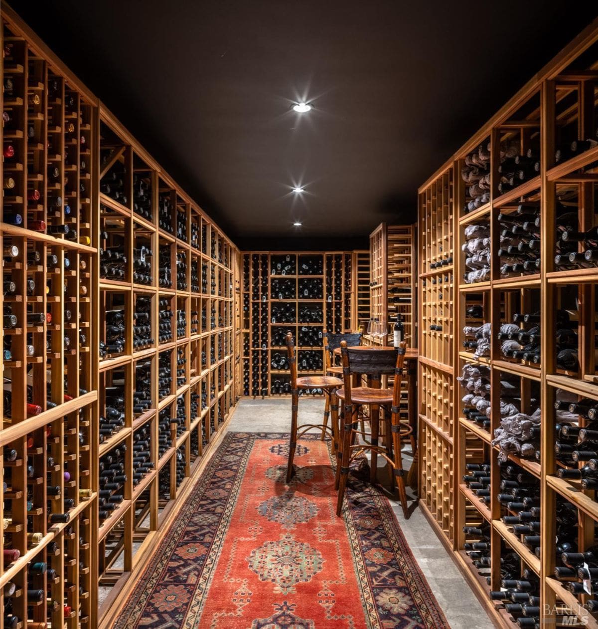 A well-organized wine cellar with wooden shelves stocked with bottles of wine. A patterned red rug and a small table with chairs create a cozy tasting area in the middle of the room.