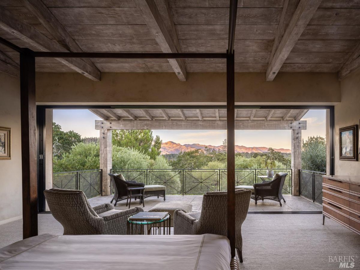 A stunning bedroom with a four-poster bed and an open view to a terrace overlooking lush greenery and mountains. The room features rustic wooden beams on the ceiling and soft neutral tones.