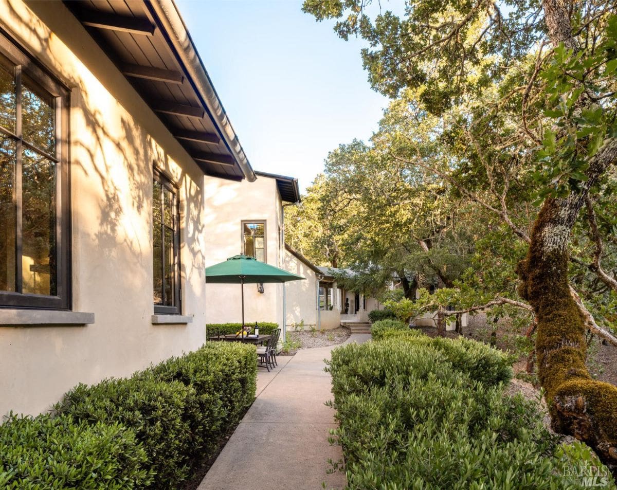 A charming side pathway lined with lush greenery and patio seating under a green umbrella. The home’s stucco walls are bathed in warm sunlight.