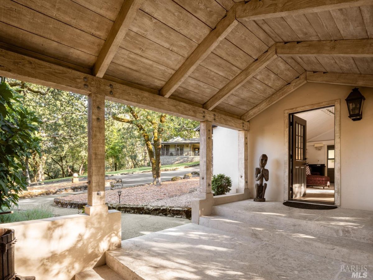 A covered porch with a rustic wooden ceiling and pillars leading to the entrance of the home. The space is surrounded by trees, creating a serene ambiance.