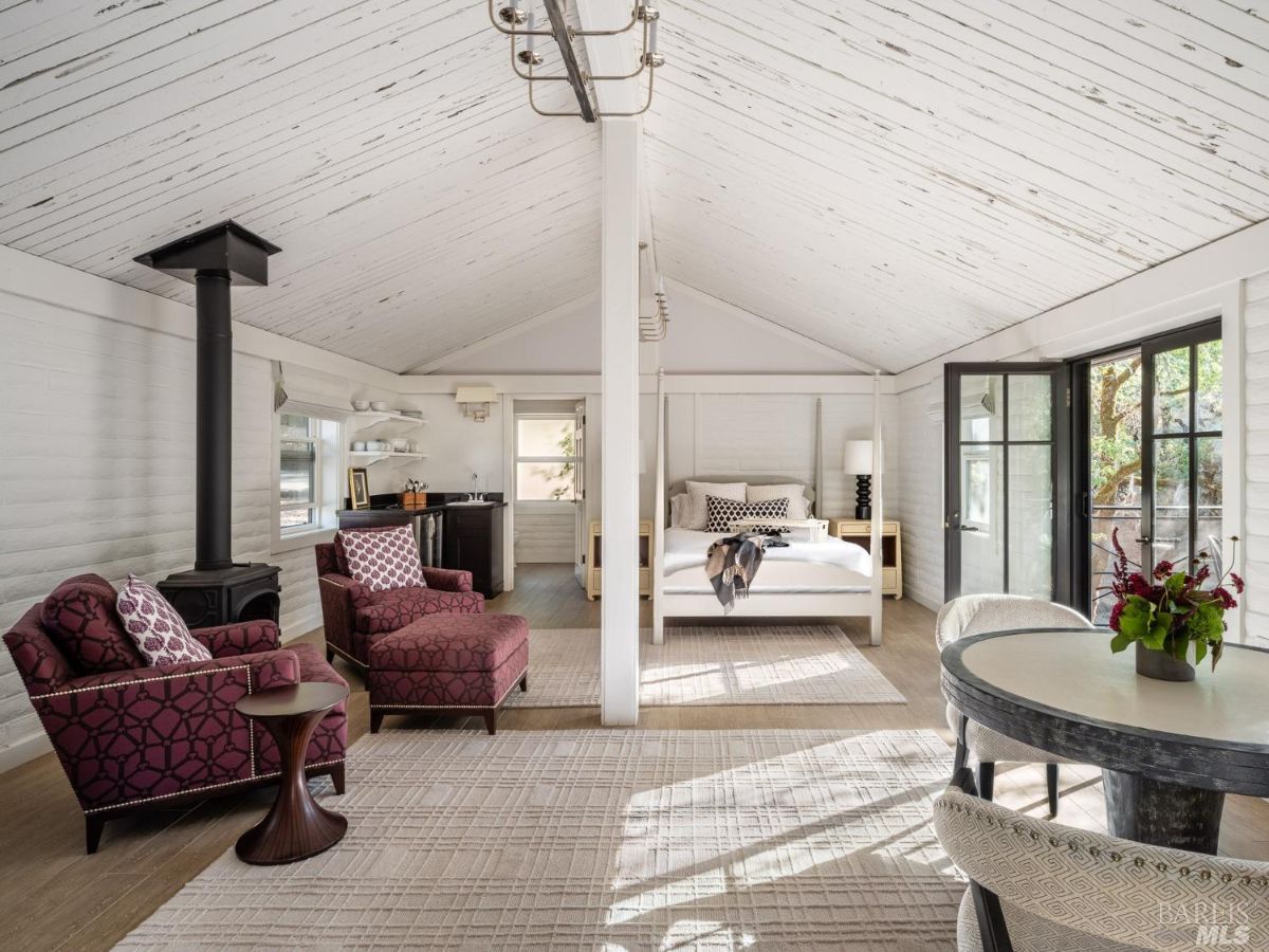 A bedroom with a four-poster bed, a cozy seating area with armchairs, and a wood-burning stove. The bright space has a minimalist yet comfortable design, with doors leading to an outdoor seating area.