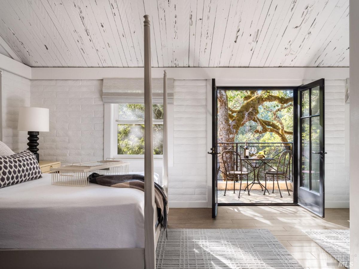  bedroom with whitewashed walls and wooden flooring, opening up to a private balcony overlooking lush greenery. The room has simple yet elegant furnishings with a focus on natural light.