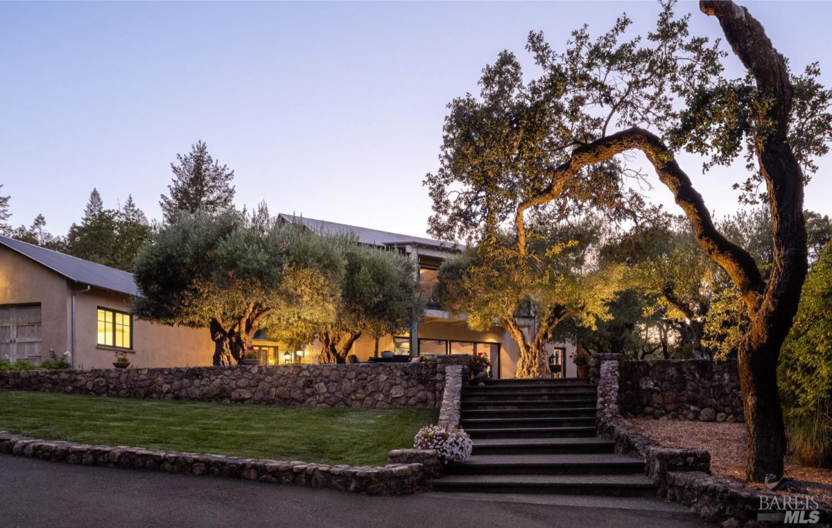 A serene evening view of a countryside estate, featuring a well-lit building surrounded by old oak trees, neatly maintained grass, and stone walls creating a picturesque entrance.