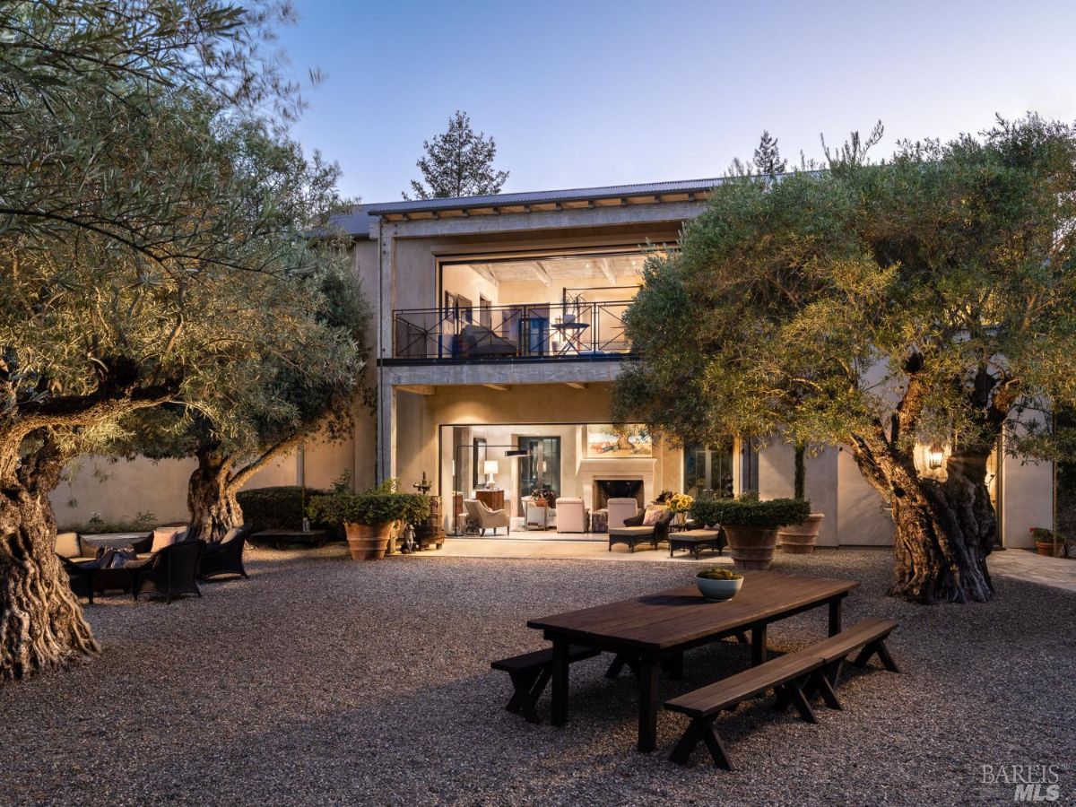 The villa's backyard features a rustic dining table and mature olive trees, offering a perfect setting for outdoor gatherings. The property is designed for both comfort and elegance.