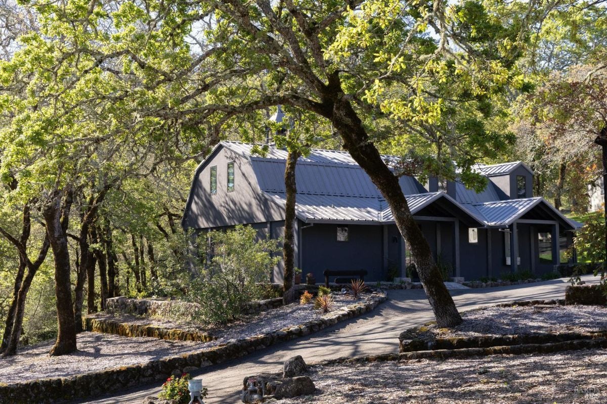 A modern gray house with a slanted metal roof surrounded by lush green trees and a landscaped path leading to the entrance. The house blends harmoniously with the natural setting, creating a serene atmosphere.