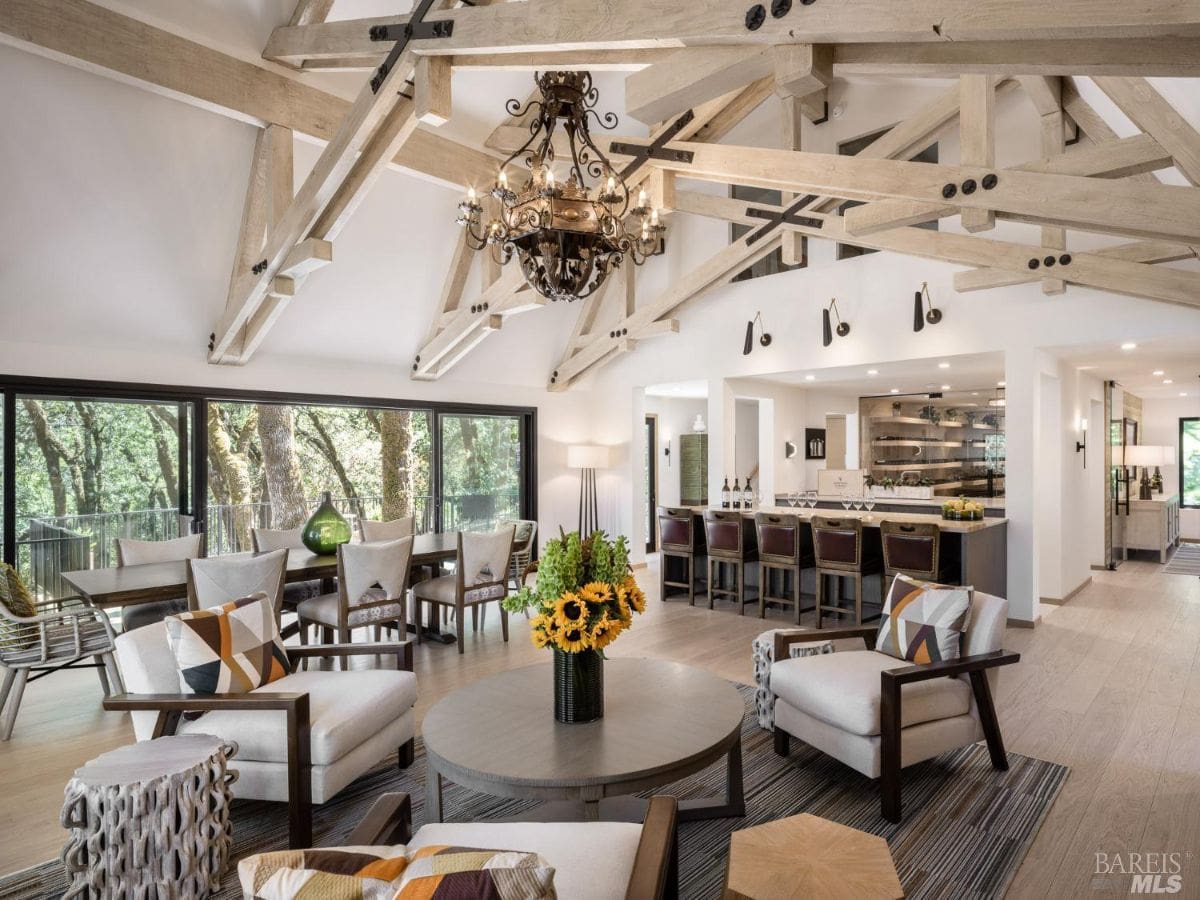  A modern and spacious dining and lounge area with a high vaulted ceiling featuring intricate wooden beams. The room includes a dining table, a bar, and large windows offering forest views.