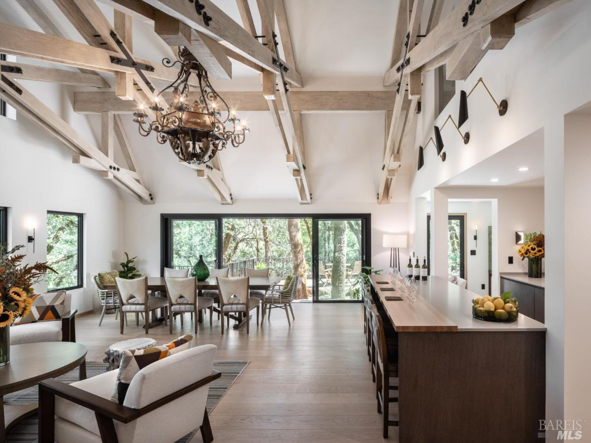 A sophisticated dining and bar area with an open layout, wooden beams, and a chandelier. The large windows allow natural light to illuminate the elegant decor and furniture.