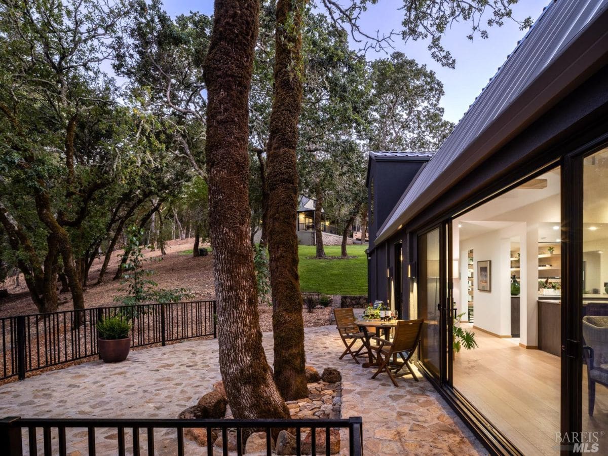 A cozy patio area with stone flooring surrounded by oak trees. Sliding glass doors connect the patio to the interior, offering a seamless indoor-outdoor