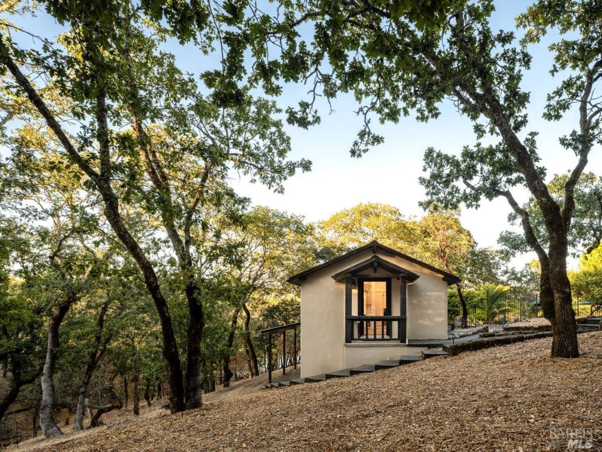 A charming small house with a stucco finish perched on a gentle slope amidst a wooded area. A pathway leads to the entrance, surrounded by natural terrain.