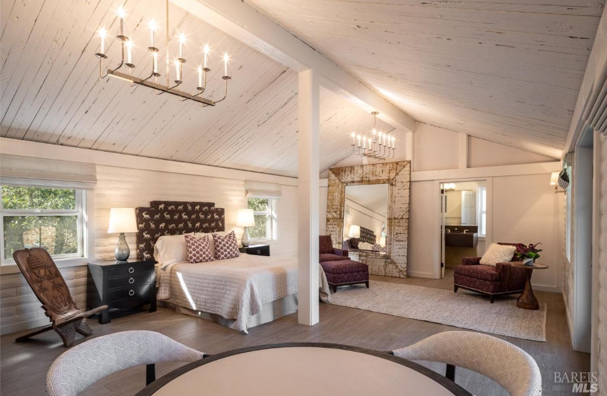  A spacious bedroom with a vaulted, whitewashed ceiling and exposed beams. The design includes a chandelier, patterned armchairs, and a large decorative mirror for a luxurious, rustic charm.