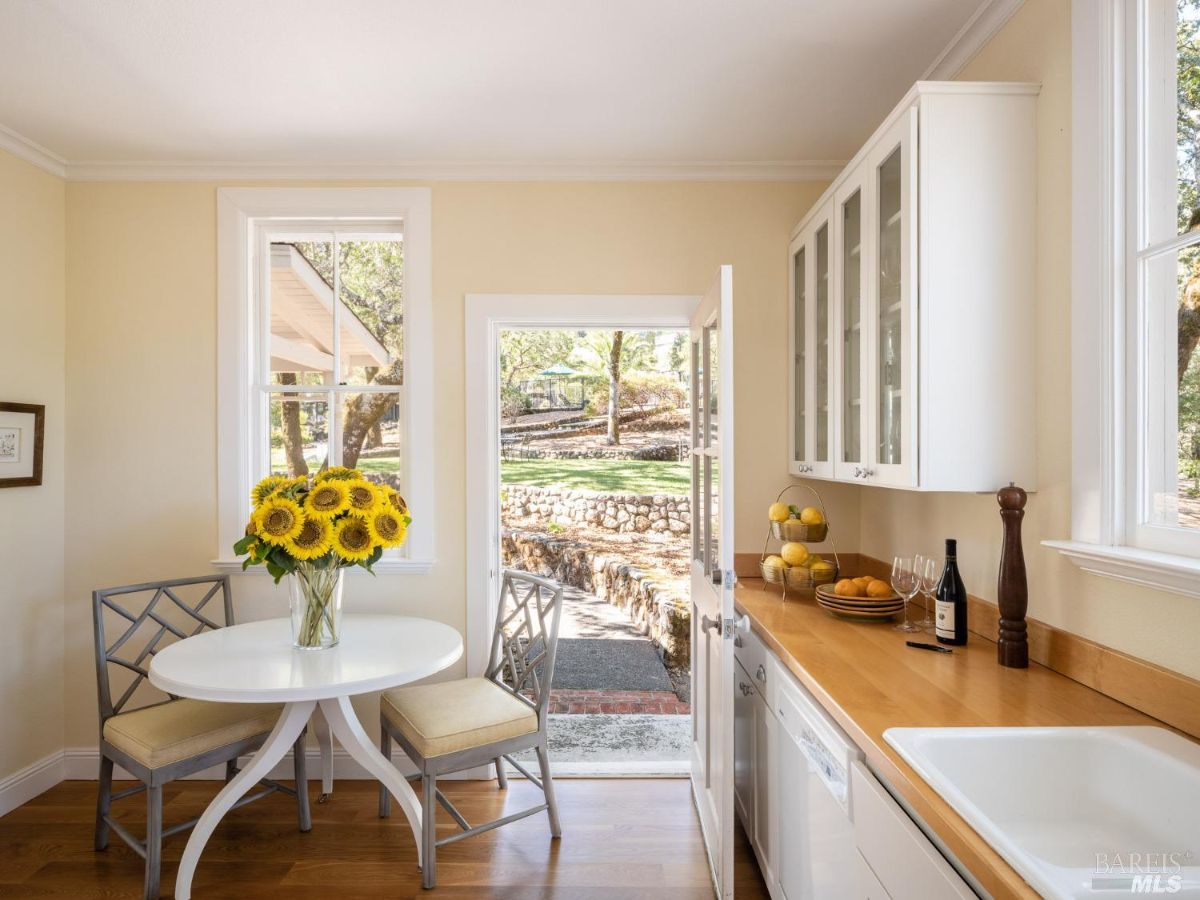 A cozy breakfast nook with a small round table and chairs, decorated with a vase of sunflowers. The bright space is complemented by large windows and a door leading outside, creating a welcoming ambiance.