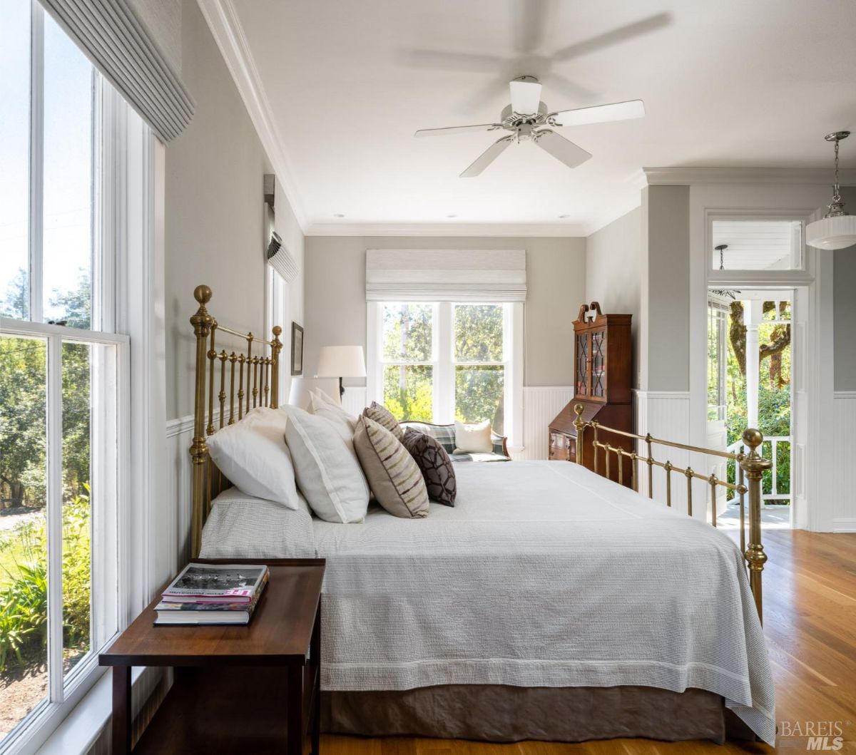  This bedroom features a brass bed frame, neutral bedding, and ample natural light from large windows. A side table with books and an armchair adds to the room's cozy, classic feel.