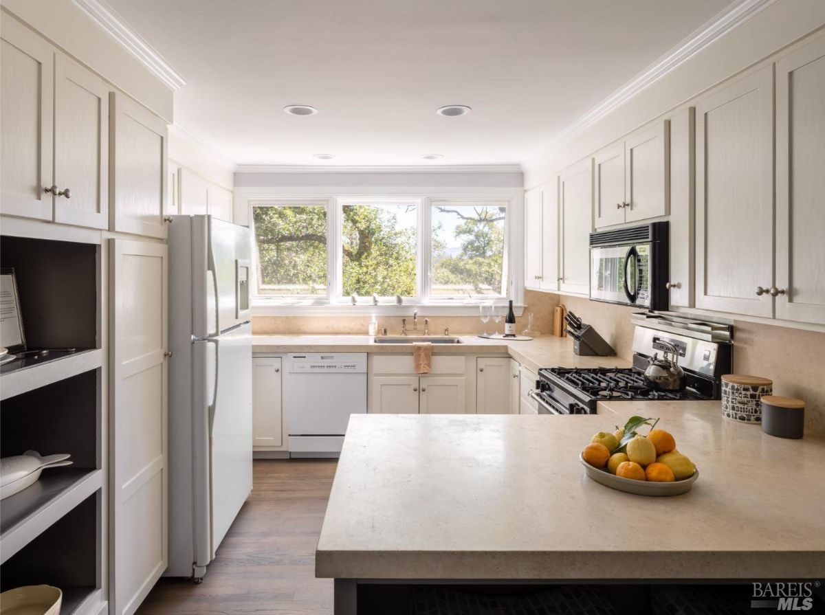 This kitchen has a clean and bright design with white cabinetry and countertops. Large windows provide ample natural light, overlooking a green landscape. The setup includes modern appliances, such as a stainless-steel stove, refrigerator, and dishwasher, and features an island in the center for additional workspace.