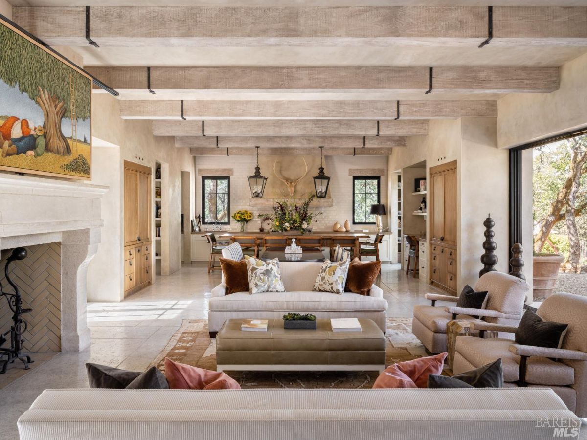 A rustic yet sophisticated living room featuring exposed wooden beams, a stone fireplace, and luxurious seating. The room is complemented by warm, neutral tones and a classic chandelier.