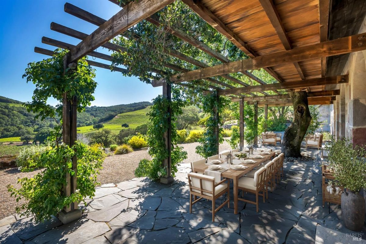An outdoor dining area on a stone patio under a wooden pergola covered with green climbing plants. A long wooden dining table with white chairs is set with tableware and lanterns. The pergola extends over the patio, casting partial shade and integrating a tree trunk into its structure. In the background, there are rolling green hills, trees, and a gravel path lined with plants. Additional seating areas with wooden benches and chairs are visible further down the patio.