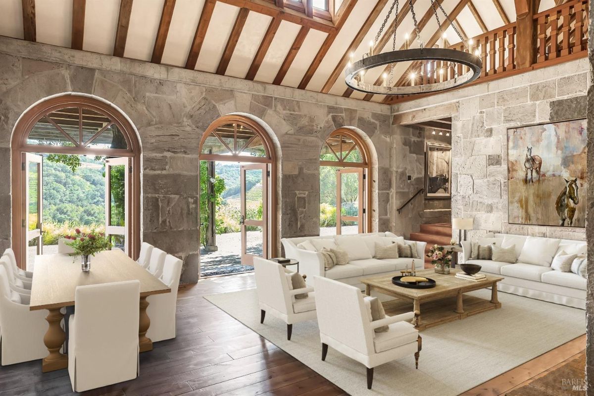 Large room with stone walls, wooden beams on the ceiling, and arched doors leading to the outdoors. The space includes a seating area with white sofas and chairs arranged around a wooden coffee table on a light-colored rug. A wooden dining table with white chairs is positioned near the doors, with a vase of flowers on the table. A large circular chandelier hangs from the ceiling, and a painting of horses is mounted on the stone wall. The arched doors provide a view of the surrounding greenery and allow natural light into the room.