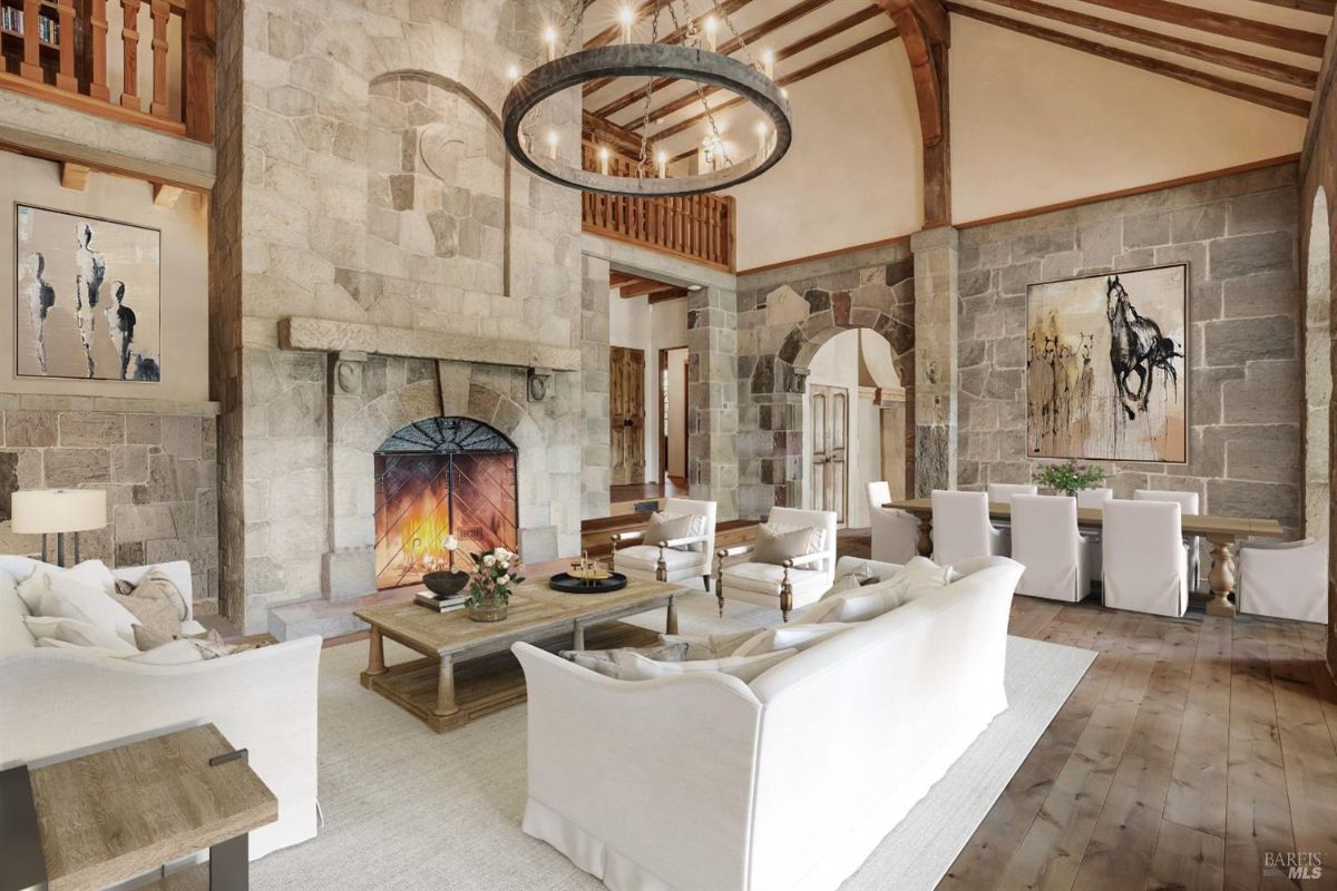 Living room with stone walls, a central stone fireplace, and wooden architectural beams. White sofas and armchairs are arranged around a rectangular wooden coffee table, which is set on a light-colored rug. A circular chandelier hangs from the ceiling, and a long wooden dining table with white chairs is visible to the right. Two paintings hang on the stone walls, one depicting abstract figures and the other a horse scene.