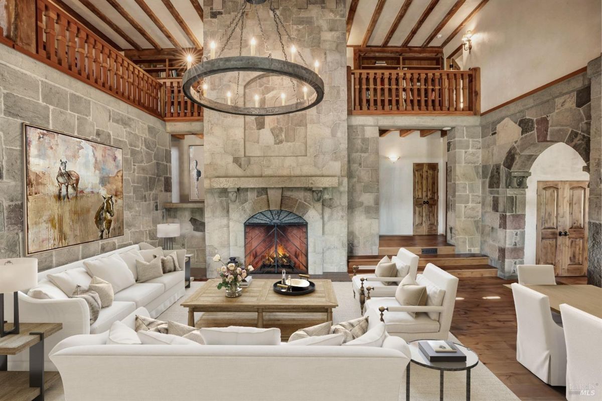 Living room with stone walls, a central fireplace, and wooden architectural details. The room has white sofas, armchairs, and a wooden coffee table arranged around the fireplace. Above the seating area, there is a circular chandelier suspended from the ceiling. A large painting featuring horses hangs on one of the stone walls. The space also includes a wooden mezzanine level with a railing, wood doors, and an archway leading to another section of the house.