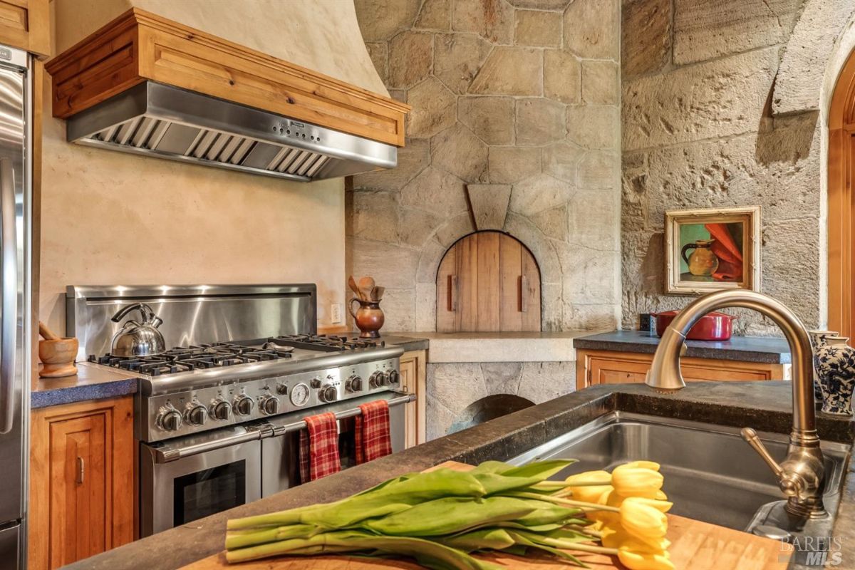 A kitchen with a stainless steel gas stove and range hood mounted under a wooden cabinet. A stone wall with an arched wood-fired oven is visible in the background. A kitchen counter with a sink and faucet in the foreground has yellow tulips on a cutting board. Wooden cabinets are present under the counters, and a framed painting hangs on the stone wall. A red pot sits on the counter near the oven, and utensils are placed next to the stove.