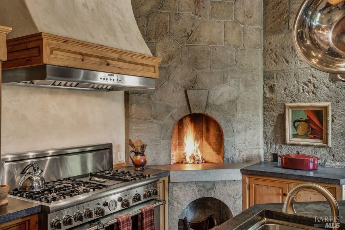 A kitchen with a large stainless steel gas stove and an overhead range hood with wooden trim. To the right, there is a stone fireplace with a fire burning inside. A countertop with wooden cabinets below is visible in the foreground, with a red pot and framed artwork resting on the surface. A small jug and utensils are placed near the stove. The walls are made of stone, and the sink faucet is visible in the bottom right corner.