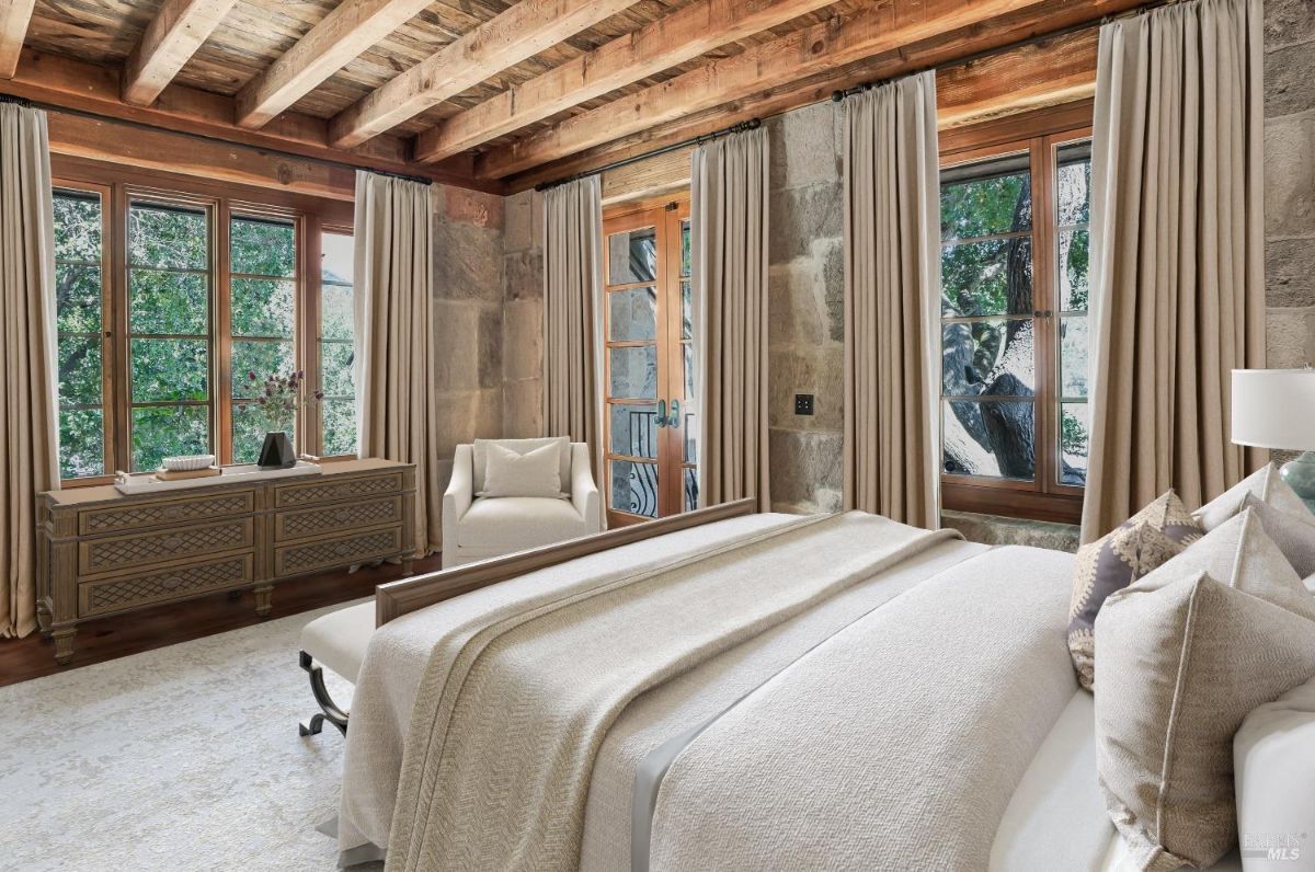 Bedroom with a large bed covered in neutral bedding and decorative pillows. A window and glass door with long curtains allow natural light to enter the room, with trees visible outside. A wooden dresser and a white armchair are placed near the windows, and a patterned area rug covers part of the floor.