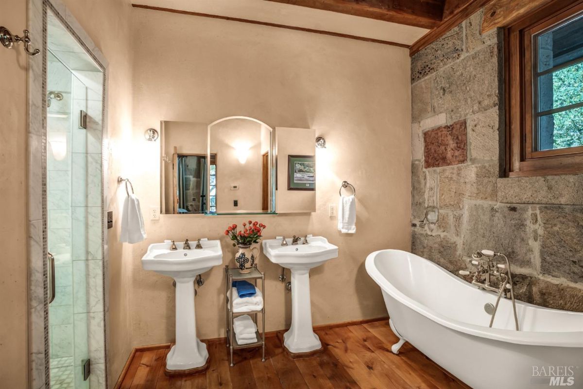 Bathroom with two pedestal sinks side by side, each with its own mirror and wall-mounted light. A freestanding white bathtub with vintage-style fixtures is placed against a stone wall. A glass-enclosed shower is on the left side, and a small shelving unit with towels and a vase of flowers is between the sinks.