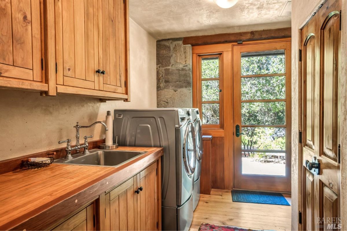A laundry room with wooden cabinets above and below a countertop with a built-in sink. Two large front-loading washer and dryer units are placed side by side near the door. A paper towel holder and small basket are situated on the countertop. A wooden-framed glass door leads outside, showing trees and greenery. The floor is made of wood, and a small rug is placed near the door.