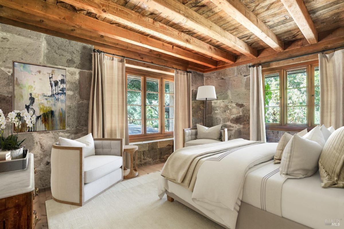 Bedroom with a large bed covered in light-colored bedding and multiple pillows. A cushioned chair with a wooden frame is placed next to a small side table, with a floor lamp nearby. The room has exposed wooden beams on the ceiling, stone walls, large windows with curtains, and a framed painting hanging on one of the walls.