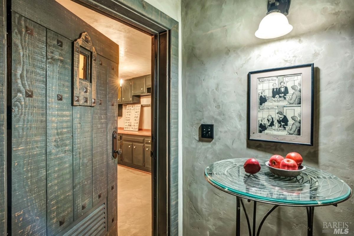 A wooden door with iron details partially open, revealing a kitchen with dark cabinetry inside. On the right, there is a small round glass-top table holding a bowl with red apples. A framed comic-style artwork hangs on the textured wall above the table, and a wall-mounted light fixture illuminates the area.