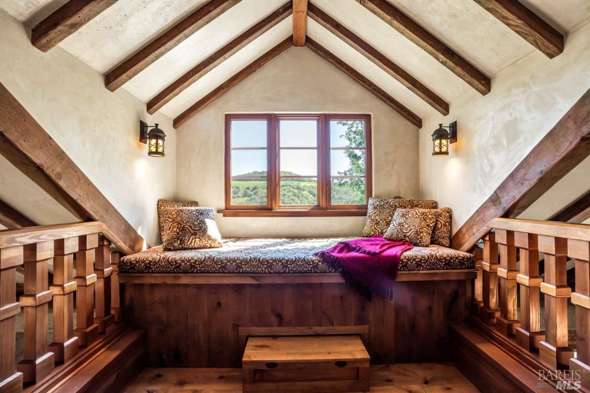 Small loft space with a built-in wooden bench under a window that has a view of rolling hills and trees. The bench has patterned cushions, two decorative pillows, and a red throw blanket. Wooden beams on the ceiling and railing frame the space, and two wall-mounted lanterns provide lighting.