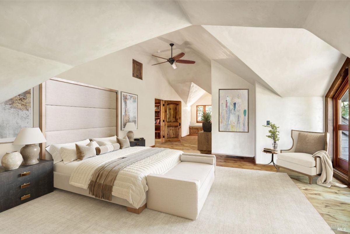 Bedroom with a large bed featuring a padded headboard, bedding, and decorative pillows. There are two dark nightstands on either side of the bed, each holding lamps and decorative items. A light-colored rug covers the floor, and a cushioned bench is placed at the foot of the bed. A separate seating area near a window includes a chair, small side table, and a potted plant. The background shows an arched wooden door leading to another space, with wooden flooring and built-in shelves visible.