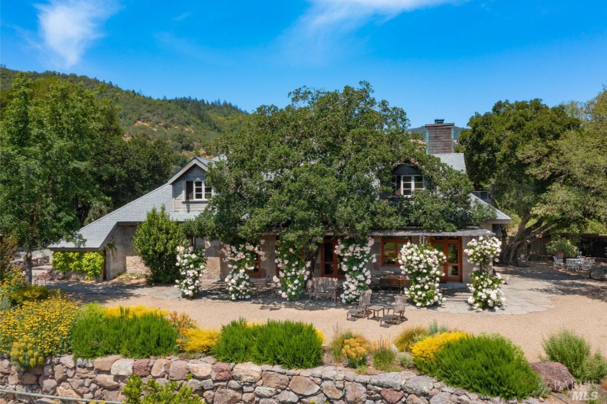 The house nestled in a landscape of trees and hills. The house is a light-colored structure with a dark roof, and surrounded by a garden with flowering bushes.