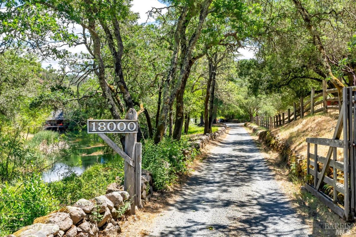 A gravel driveway lined with trees and bordered by a wooden fence on the right and a stone wall on the left. A wooden signpost with the number "18000" is visible at the entrance. To the left, a pond surrounded by greenery reflects the trees, while the driveway curves into the shaded wooded area ahead.