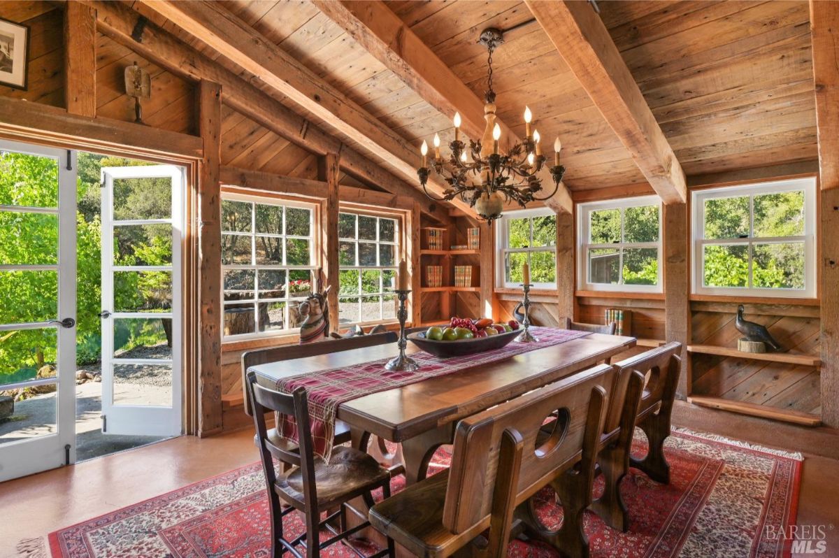 Dining room with wooden walls, ceiling beams, and large windows allowing natural light. A wooden dining table with a red plaid table runner is surrounded by matching wooden chairs. A chandelier hangs from the ceiling above the table, and a red patterned rug covers the floor. Built-in shelves on the back wall hold books and decorative items, including two bird figurines. Double doors open to an outdoor area with greenery visible outside.