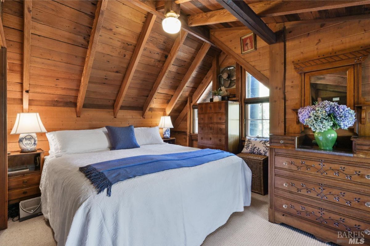 Bedroom with wooden walls and exposed beams on the ceiling. A bed with white bedding and blue accents is placed against the wall, with two lamps on small nightstands beside it. A wooden dresser with a mirror and a vase of flowers is on the right, and a chest of drawers is positioned near a window in the background.