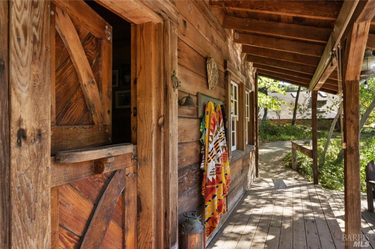 Wooden porch with a half-open rustic wooden door leading into the structure. Two colorful blankets hang beside the door, and a small bell is mounted on the wall. The porch is covered with wooden beams, and the floorboards extend into a pathway surrounded by trees and greenery.