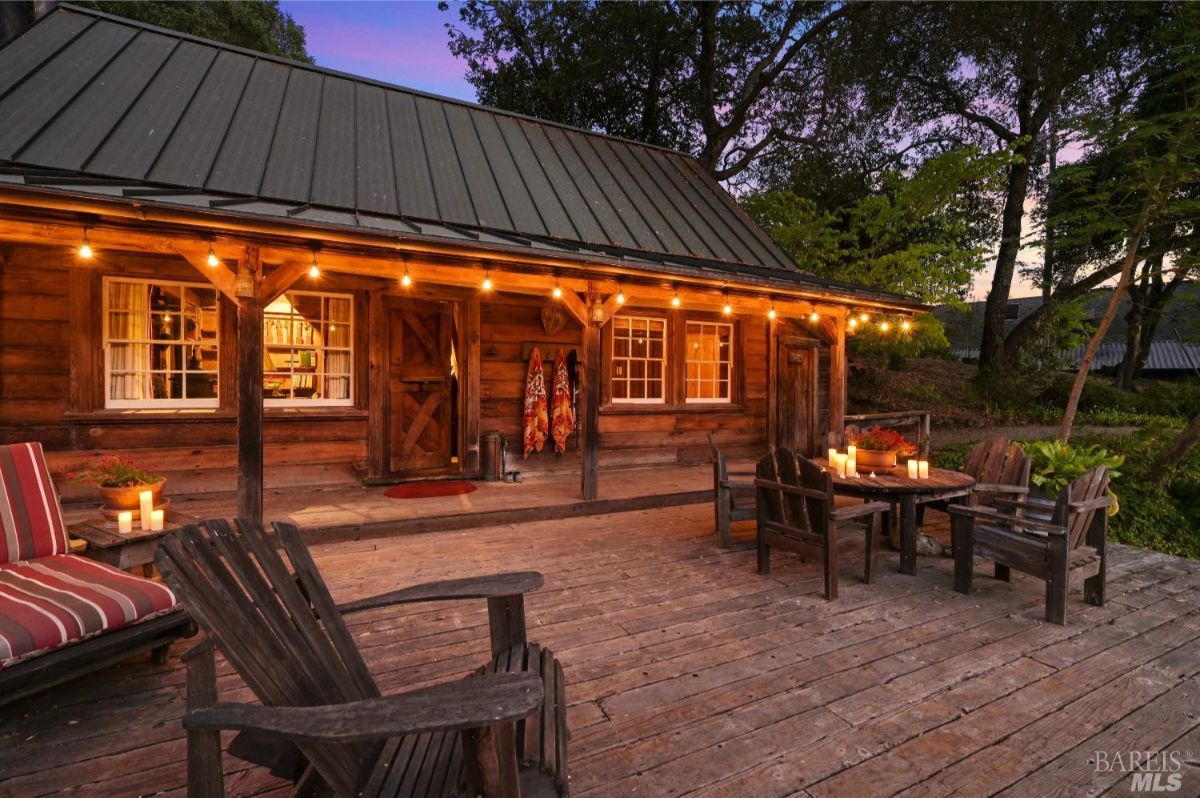 Wooden cabin with a covered porch lit by string lights at dusk. The porch features several Adirondack chairs, a wooden table with candles, and a bench with red-striped cushions. Trees and plants surround the cabin, and warm light can be seen glowing through the windows.