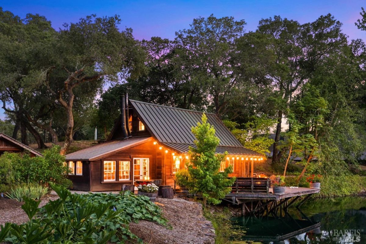 Wooden cabin with a metal roof surrounded by large trees and lush greenery. The cabin has a wrap-around deck with string lights illuminating the outdoor seating area. A small pond or reflective body of water is visible at the base of the deck, reflecting the lights and surrounding trees.