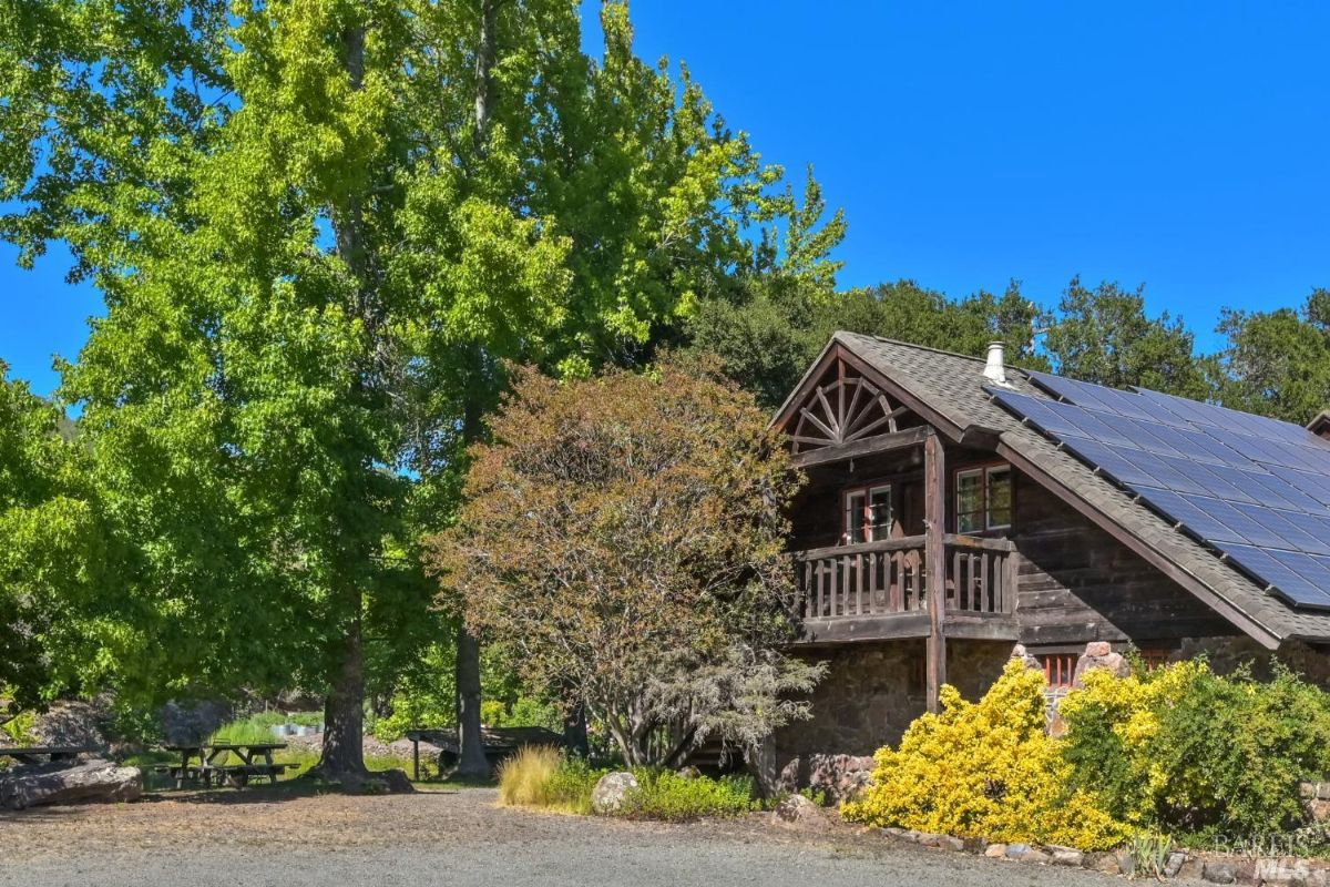 Rustic wooden building with a steep roof that has solar panels installed on it. A small balcony with wooden railings is visible on the building's second level. The area around the structure includes trees, a bush with yellow foliage, a gravel driveway, and picnic tables in the background.
